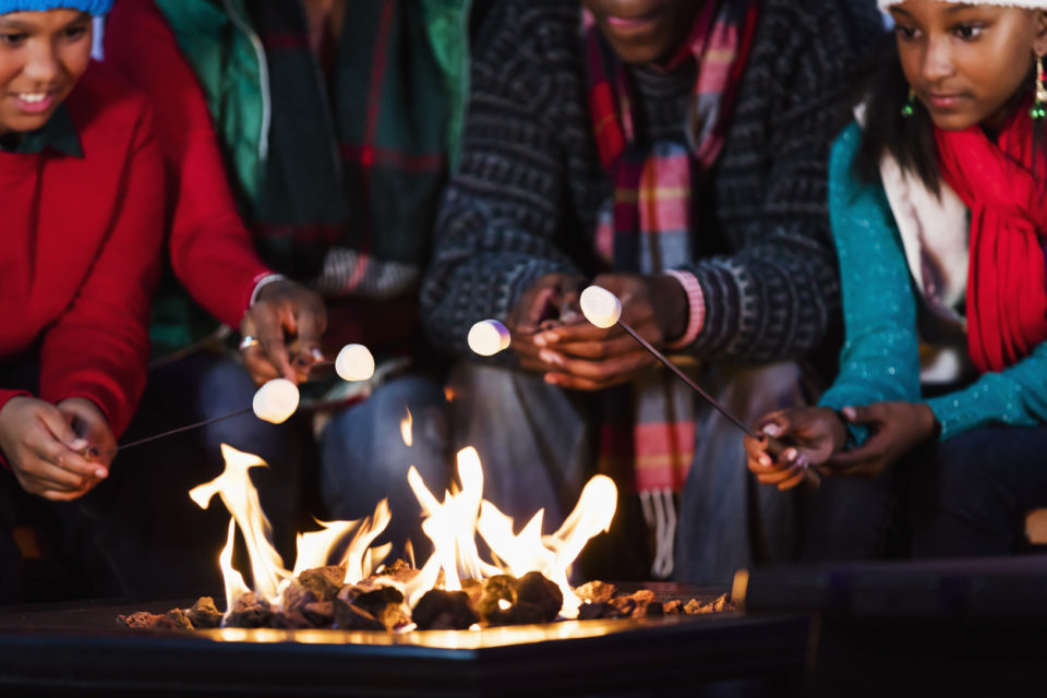 Close-up of a family with two children sitting together around a fire pit, roasting marshmallows. They are wearing winter scarves and sweaters.