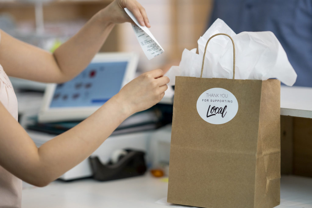 Retail worker placing debit receipt in a paper shopping bag with a "Thank You for Supporting Local" sticker on it.