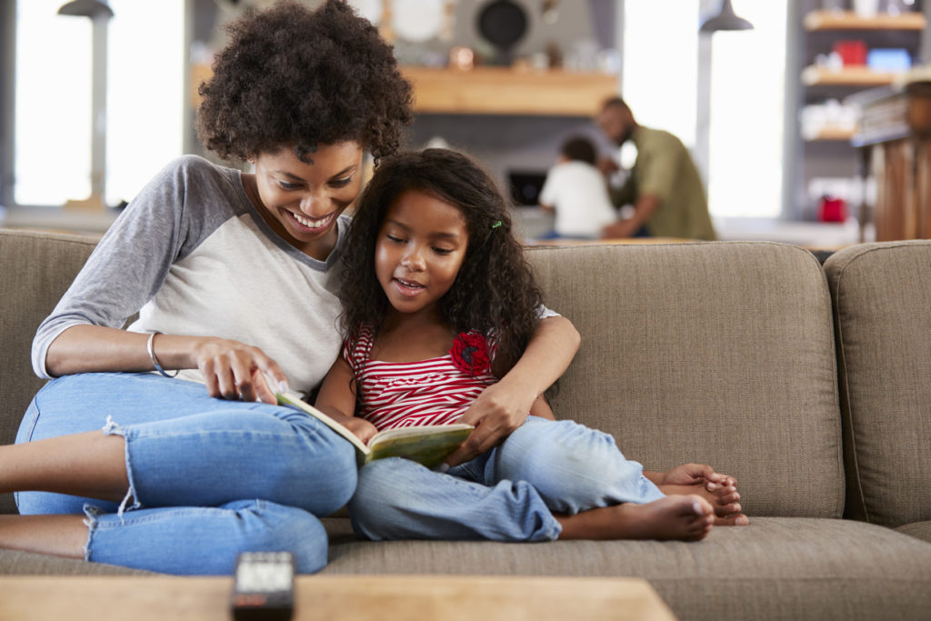 Mother And Daughter Sit On Sofa In Lounge Reading Book Together