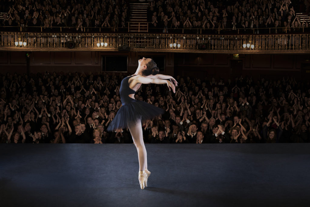 A ballerina performs on stage in front of a packed audience.